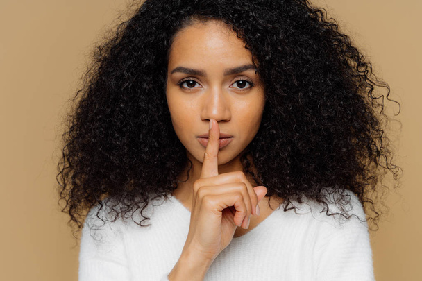 Image of serious confident dark skinned woman makes shush gesture, keeps fore finger over lips, tells secret information, asks not spread rumors, dressed in casual outfit, isolated on beige background - Photo, Image