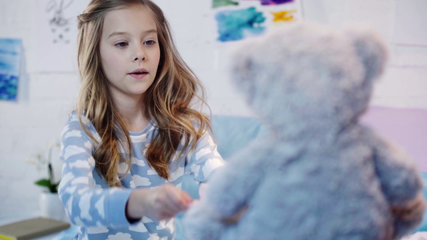 aranyos preteen gyermek pizsamában a mackó közelében ülve, és játszik a játék edények a hálószobában - Felvétel, videó