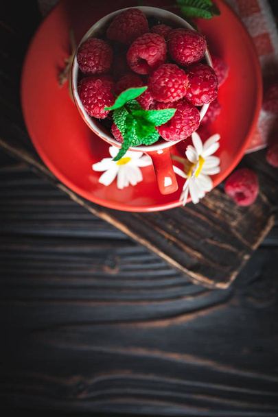 Raspberry in a red cup with chamomile and leaves on a dark background. Summer and healthy food concept. Selective focus. Background with copy space. Flat lay. - Фото, зображення