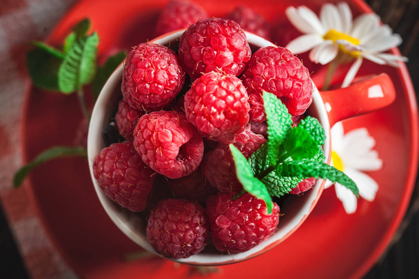 Raspberry in a red cup with chamomile and leaves on a dark background. Summer and healthy food concept. Selective focus. Flat lay. - Фото, изображение