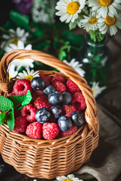 Raspberries and blueberries in a basket with chamomile and leaves on a dark background. Summer and healthy food concept. Selective focus. - Foto, afbeelding
