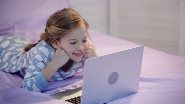 preteen kid in pajamas lying on bed, propping face with hand and using laptop with smile - Filmmaterial, Video