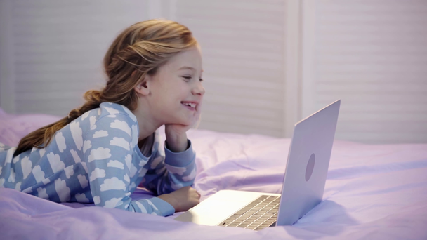 niño preadolescente en pijama acostado en la cama mientras escribe en el teclado del ordenador portátil, apuntalando la cara con la mano, sonriendo y señalando con el dedo en la pantalla
 - Imágenes, Vídeo