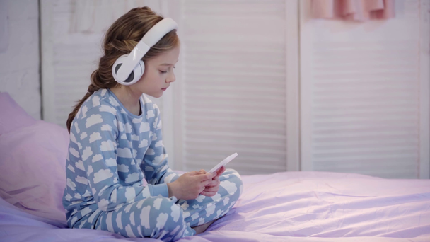 gyermek pizsama ül az ágyban, zenét hallgat fejhallgató és smartphone segítségével - Felvétel, videó