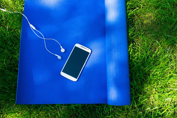 A mat for yoga and pilates, a phone with headphones and on green grass, concepts of training and sports in nature - Photo, Image