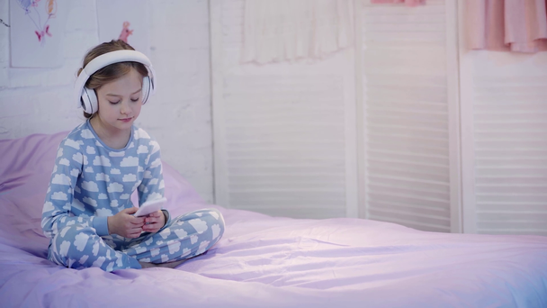 child in pajamas sitting on bed, listening music in headphones and using smartphone - Footage, Video