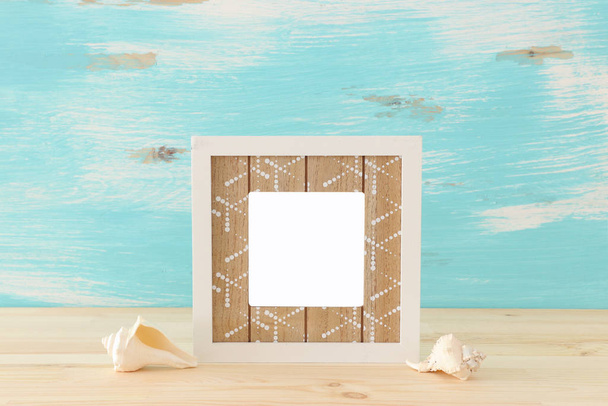 blank white photo frame and sea shells over wooden table and pastel blue background. Ready for photography montage - Fotó, kép