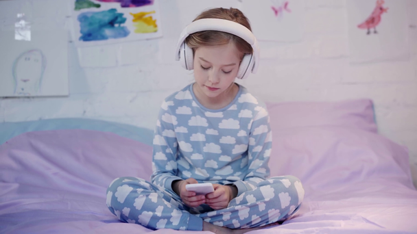 child in pajamas sitting on bed, listening music in headphones and using smartphone - Felvétel, videó