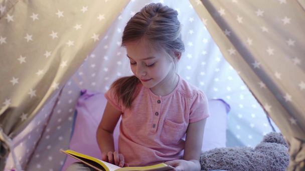 smiling focused preteen kid sitting in wigwam and reading book - Footage, Video
