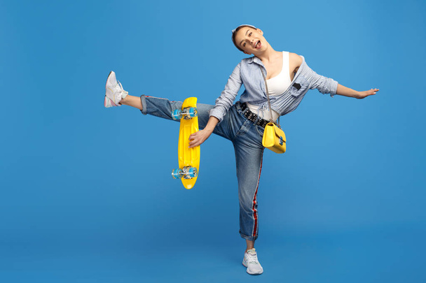 Full length photo of happy young woman with yellow penny or skateboard dancing over blue background. - Foto, imagen