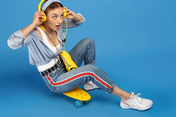 Full length photo of happy carefree young woman with yellow headphones sitting on penny or skateboard over blue background. - Fotoğraf, Görsel