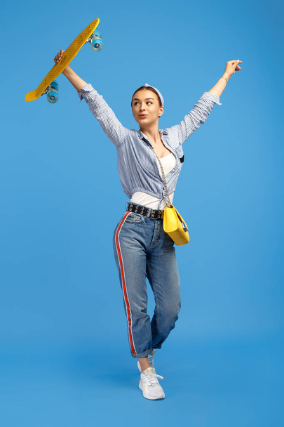 Full length photo of happy young woman with yellow penny or skateboard dancing over blue background. - Photo, image