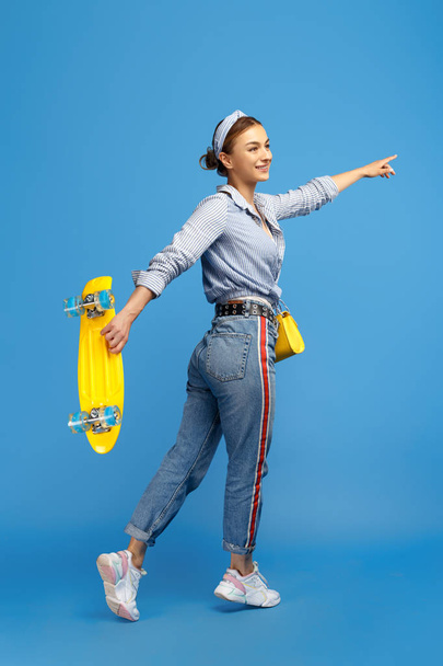 Full length photo of happy young woman with yellow penny or skateboard dancing over blue background. - Fotó, kép