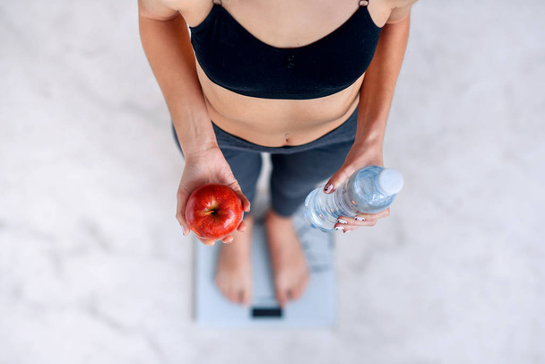 Sporty woman with a perfect body measuring body weight on electronic scales and holding a bottle of water and a red apple. Healthy low calorie meals, fruits. Diet, a healthy lifestyle. - Фото, зображення