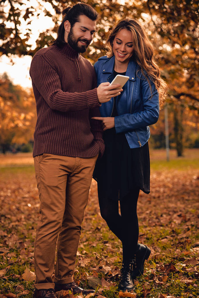 Νεαρό ζευγάρι χρησιμοποιώντας κινητό στο φθινόπωρο χρωματιστά πάρκο. - Φωτογραφία, εικόνα