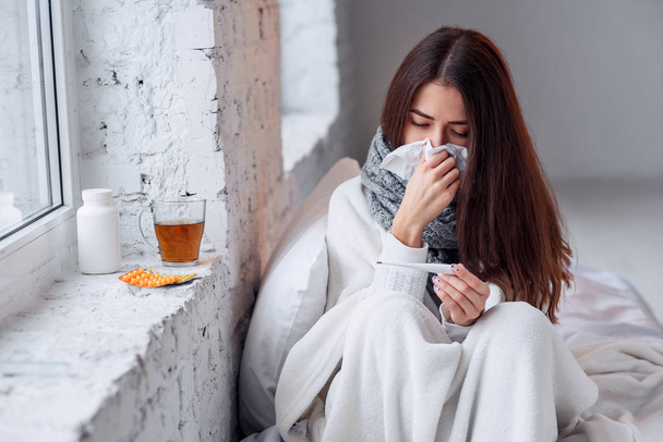 Sick woman caught cold, feeling illness and sneezing in paper wipe. Closeup of beautiful unhealthy girl covered in blanket wiping nose and looking at thermometer. Healthcare concept. - Photo, Image