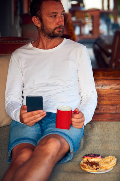 Mann sitzt auf einem Terrassensofa und trinkt Kaffee / Tee - Foto, Bild