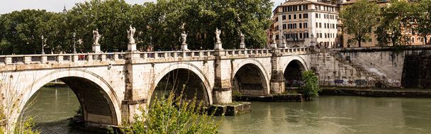 ROME, ITALY - JUNE 28, 2019: panoramic shot of river Tiber and people walking on old bridge in sunny day - Foto, Bild
