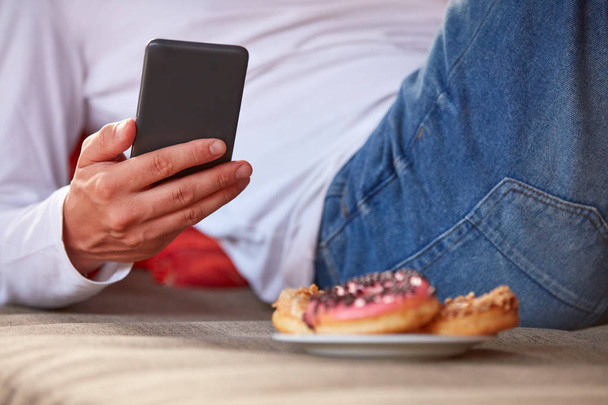 Homem sentado em um sofá terraço e comer donuts enquanto estiver usando celular
 - Foto, Imagem