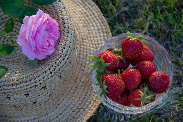 熟したイチゴクリスタルの花瓶、お茶のバラはわらの上にバラの女性の帽子は、庭の緑豊かな草の上に横たわっています. - 写真・画像