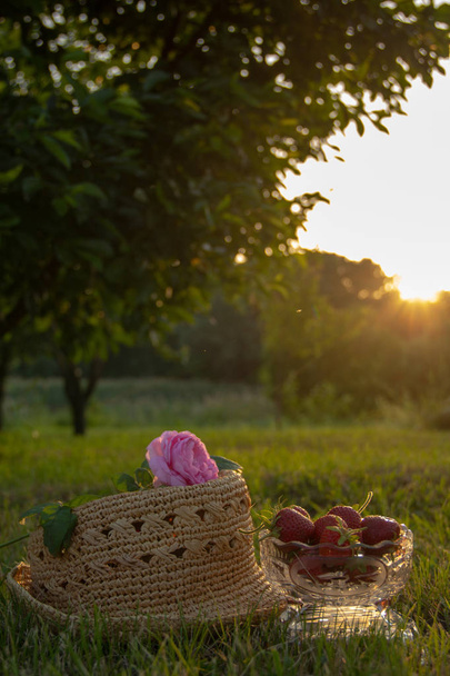 熟したイチゴの結晶花瓶、わらの上の茶のバラは庭の緑の草の上にあり、背景には木と太陽光線があります. - 写真・画像