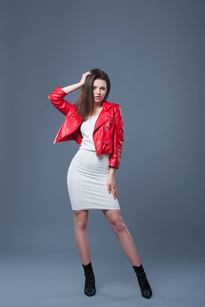 Fashionable style, fashion women's clothing, color combination. Beautiful brunette girl in white dress and red leather jacket isolated gray background - Foto, afbeelding