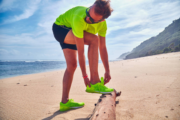 Man tying running sneakers on a tropical exotic beach. - Фото, изображение