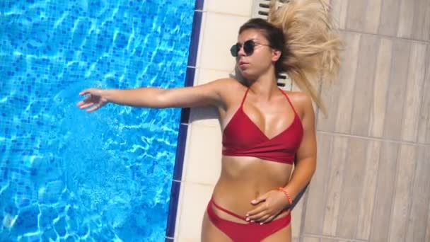 Close up of attractive tanned woman in sunglasses lying on basin edge and touching water surface. Sexy girl sunbathing near pool and enjoying rest. Concept of summer vacation. Top view Slow motion - Footage, Video