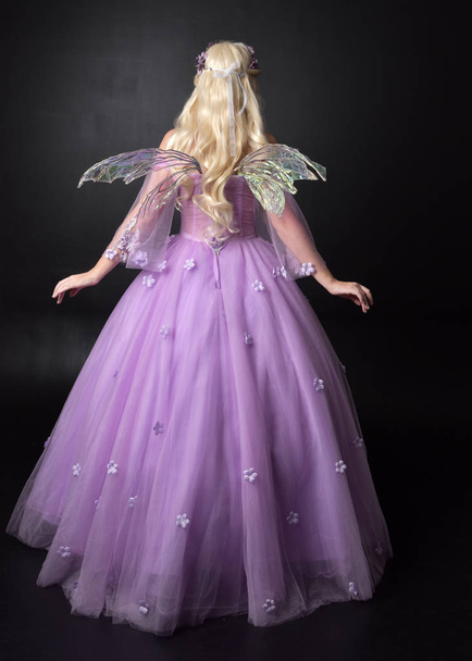 full length portrait of a blonde girl wearing a fantasy fairy inspired costume,  long purple ball gown with fairy wings,   standing pose  with back to the camera on a dark studio background. - Photo, Image