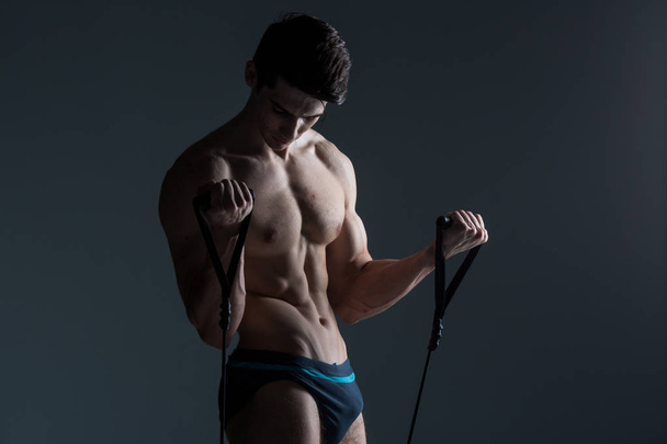 Muskularny fitness młody męski zabytkowe doskonały mięśnie sześć opakowania z ABS i nagi skrzynia. Kulturysta model pociągów z rozciągania elastyczny na ciemnym tle w studiu. Trening treningowy siłownia - Zdjęcie, obraz