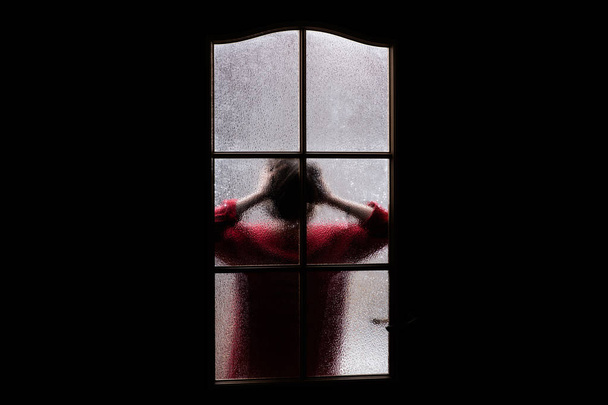 Ciemna sylwetka dziewczynki w kolorze czerwonym za szkłem. Zamknięty samotnie w pokoju za drzwiami na Halloween. Koszmar dziecka z obcymi, potworami i upiorami. Zło w domu. Wewnątrz Nawiedzony dom. Nadprzyrodzone. - Zdjęcie, obraz