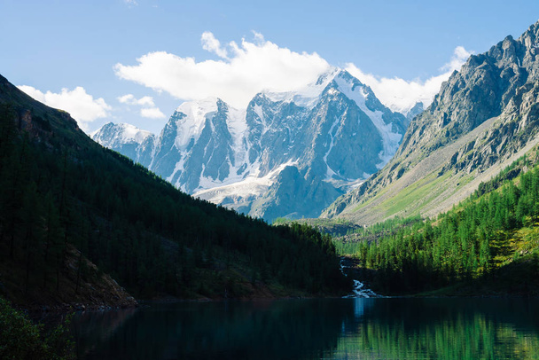 Amazing glacier under blue sky. Forest reflected on clean water of mountain lake. Huge cloud on giant wonderful snowy mountains. Rich vegetation of highlands. Atmospheric landscape of majestic nature. - Photo, Image