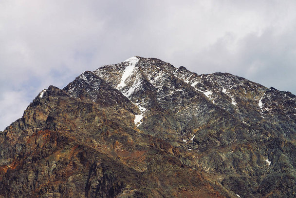 Χιονισμένη κορυφή βουνού σε συννεφιασμένο ουρανό. Βραχώδης κορυφογραμμή κάτω από σύννεφα. Συννεφιασμένη καιρός στα υψίπεδα. Ατμοσφαιρικό μινιμαλιστικό τοπίο της μεγαλειώδης φύση. - Φωτογραφία, εικόνα