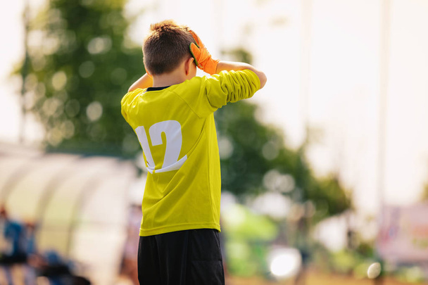Λυπημένος μικρός τερματοφύλακας ποδοσφαίρου. Απογοητευμένος αγόρι με κίτρινο τερματοφύλακα σετ αθλητικών ρούχων με τα χέρια στο κεφάλι του. Ομάδα ποδοσφαίρου νεαρών εφήβων που χάνει τον στόχο - Φωτογραφία, εικόνα