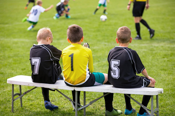 Banc de football. Jeunes footballeurs assis sur le banc de remplaçant de football. Match de football et arbitre en arrière-plan
 - Photo, image