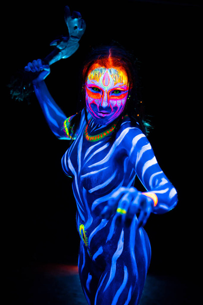 Portrait of Young naked bodyarted woman in blue glowing ultraviolet paint with tomahawk ancient prehistorical weapon. Agressive avatar warrior amazon with pigtails hairstyle - Photo, Image