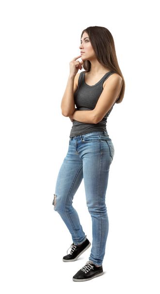 Young woman in sleeveless gray top and blue jeans standing in half turn, deep in thought, with fingers rubbing her chin, isolated on white background. - Foto, imagen