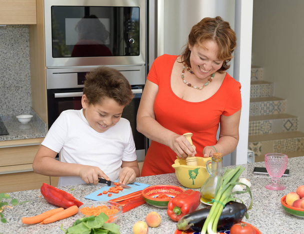 Η μητέρα και ο γιος ετοιμάζουν μεσημεριανό και χαμόγελα. Ο γιος κόβει το κόκκινο πιπέρι - Φωτογραφία, εικόνα