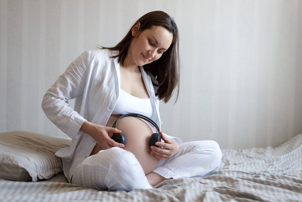 Retrato de una chica embarazada en la cama con auriculares en la barriga, audio terapia
 - Foto, Imagen