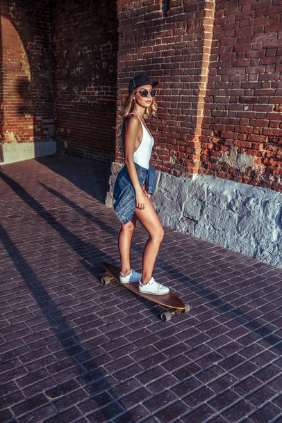 The girl in the summer rides a skateboard, rest in city at the weekend. Fashion youth style, modern bold, daring lifestyle. Concept walk rest emotions smile and lay woman in white bodysuit. - Фото, изображение