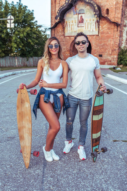 A young couple a man and a woman walk in the summer in the city at the weekend, the happy ones are smiling joking and having fun. Skateboard board longboard, casual wear, love relationship concept. - Photo, Image