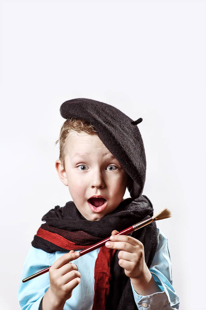 boy artist in black beret, scarf and with a brush in his mouth on a light background - Photo, Image