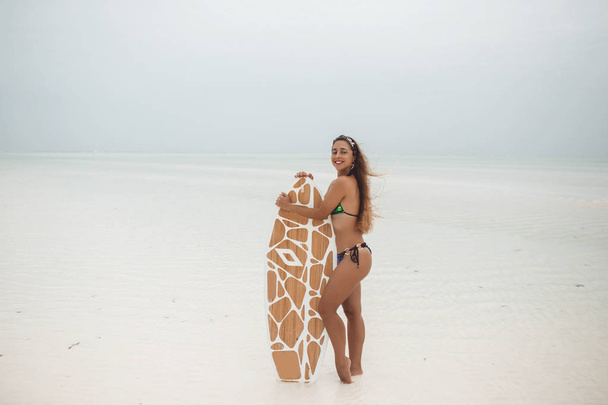 Young sexy woman with sportive tanned body, smiling and jogging with surf board on the beach in cloudly summer day during low tide in Zanzibar - Zdjęcie, obraz