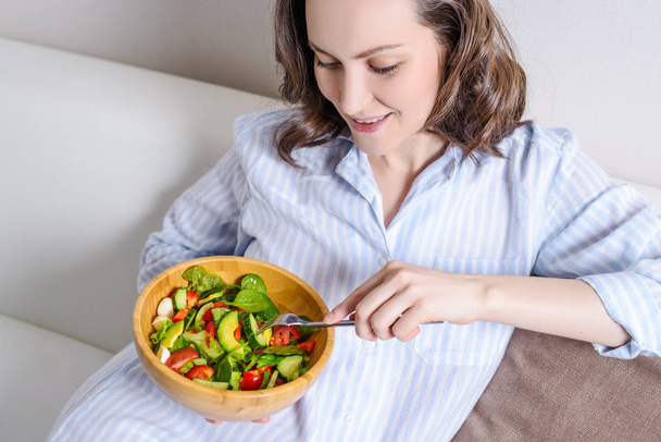 Νεαρή χαμογελαστή γυναίκα με μπλε πουκάμισο τρώγοντας φρέσκια σαλάτα λαχανικών - Φωτογραφία, εικόνα