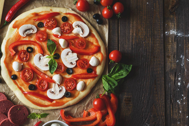 Platte lay pizza deeg met tomaten, champignons, paprika, olijven en spinazie met kopieerruimte op getextureerde houten tafel - Foto, afbeelding