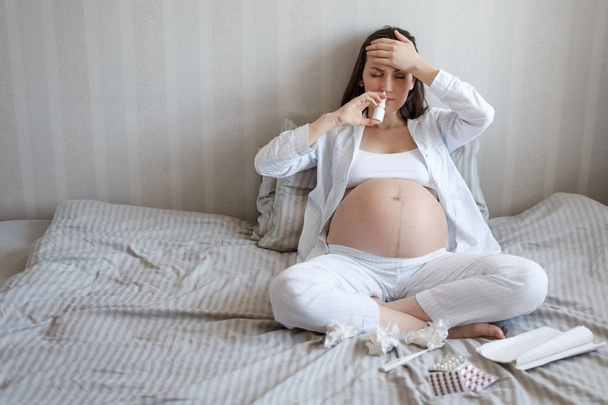Μετωπικό πορτρέτο μιας εγκύου που κάθεται στο κρεβάτι με ένα κρύο σπρέι, χάπια, χαρτομάντιλα, θερμόμετρο. κρυολογήματα κατά τη διάρκεια της εγκυμοσύνης, υγειονομική περίθαλψη - Φωτογραφία, εικόνα