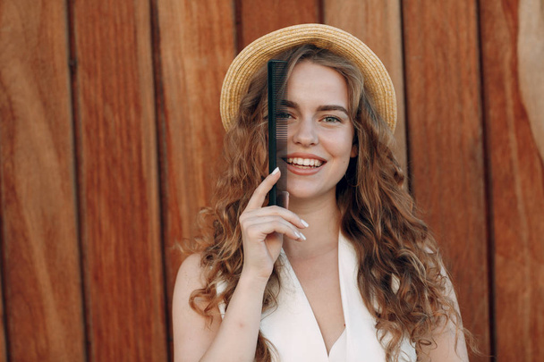 Porträt einer jungen lächelnden positiven Frau mit lockigem Haar in Strohhut mit Kamm. Haarstyling-Konzept. - Foto, Bild