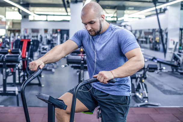 young athlete using exercise bike at the gym. Fitness male using air bike for cardio workout at crossfit gym and listenin music. dressed in sportswear - Photo, Image