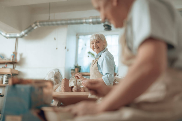 Waist up of happy elderly craftswoman painting earthenware in potters studio - Foto, imagen
