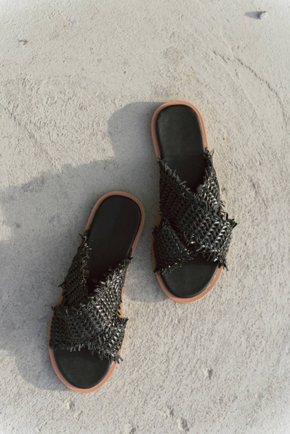 zwarte rieten schoenen met platte zool. Stijlvolle zomer leien zonder poten. Modieuze vrouwelijke slippers op grijs asfalt met een schaduw op het. - Foto, afbeelding